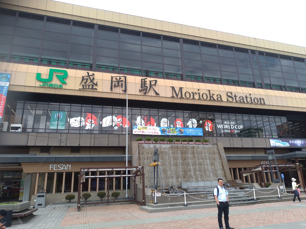 盛岡駅東口。ガラス張りの高架ホームにE5系新幹線が停車している。駅前に立っているのは佐藤先生